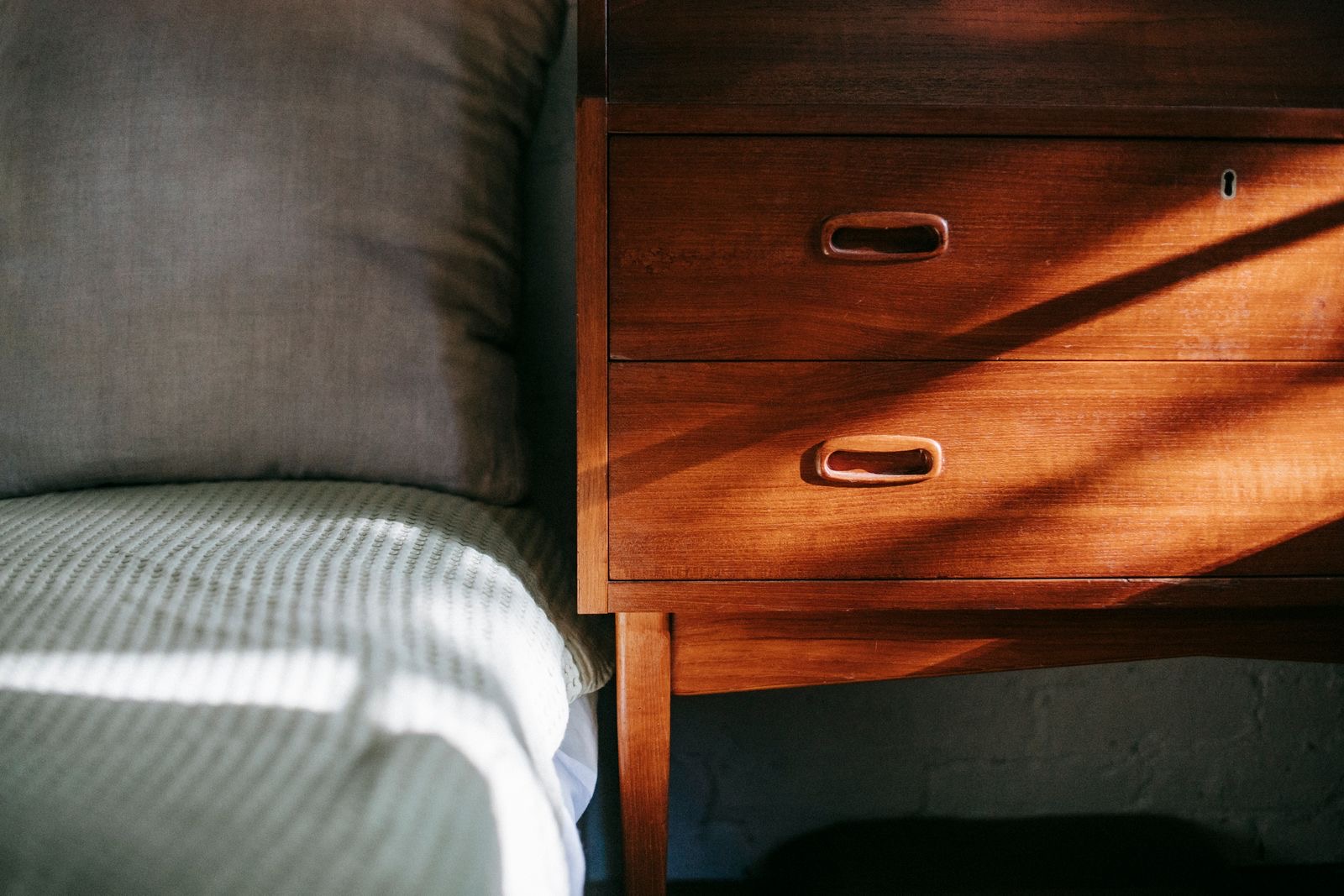 edge of a bed with a gray pillow beside a wooden nightstand with two drawers in morning sunlight