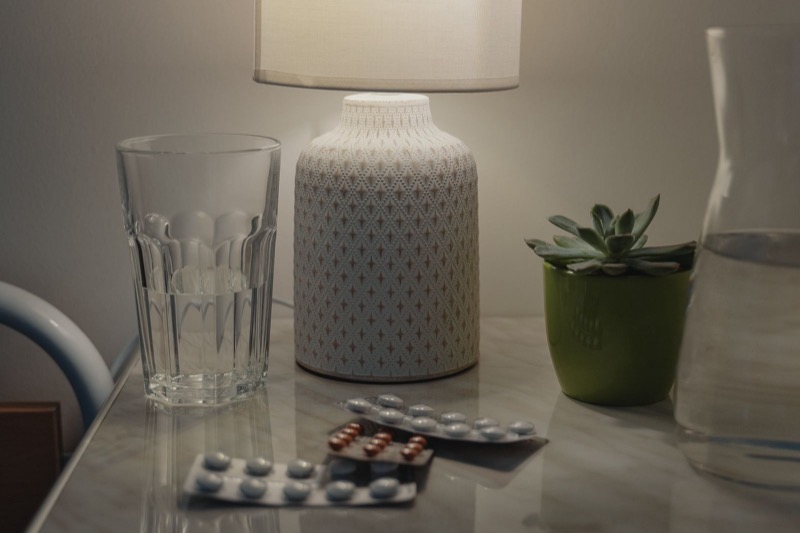 lit table lamp, a glass of water and two blister packs of pills on a marble nightstand
