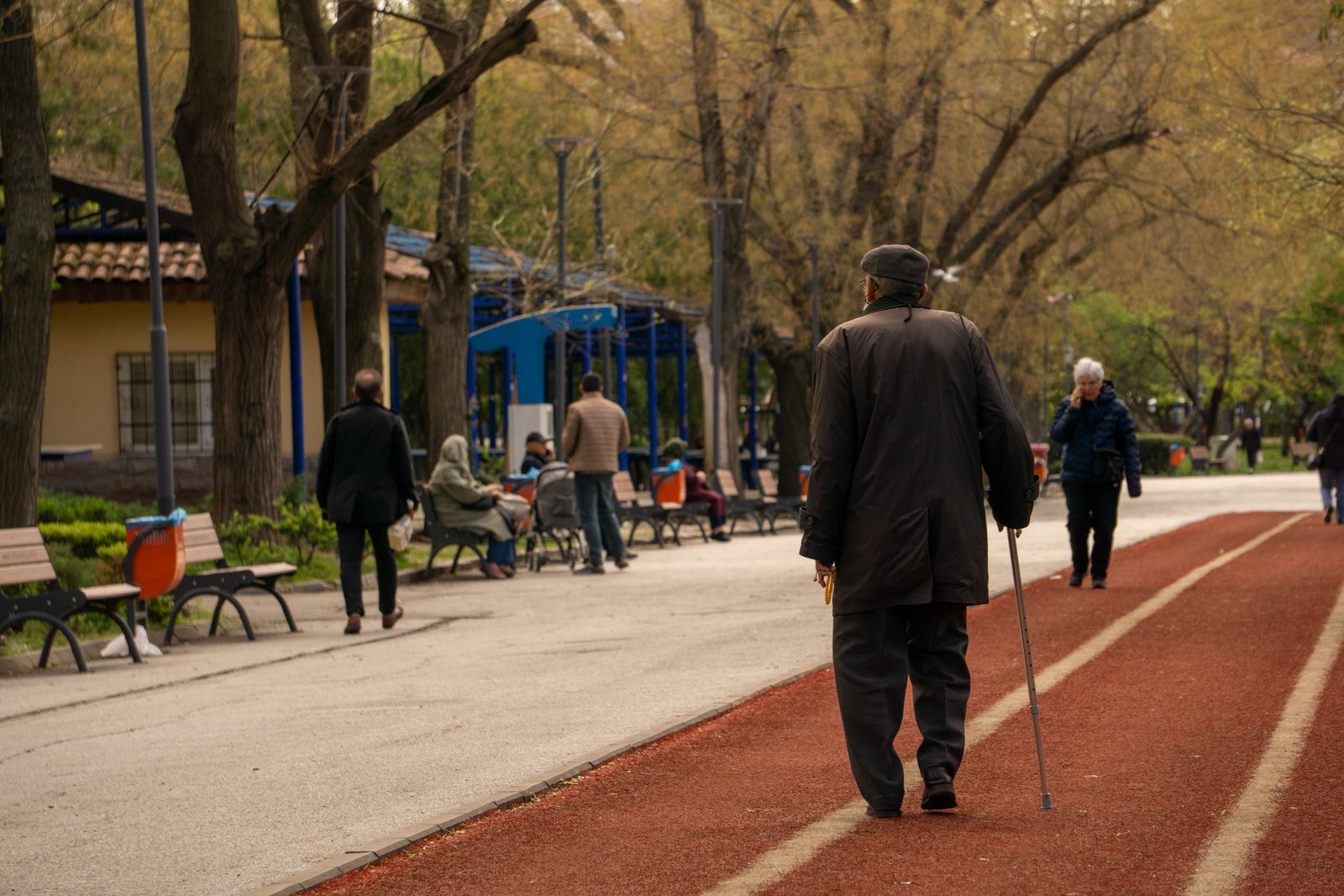 older man in a dark coat and cap walking away with a cane along a red path in a tree-lined park