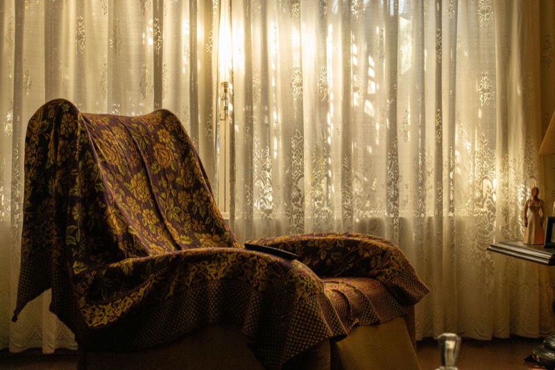 armchair covered with a patterned throw blanket in front of sunlit lace curtains