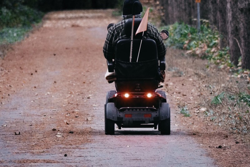 person riding a four-wheel mobility scooter away along a gravel path with a flag on the back and tail lights on