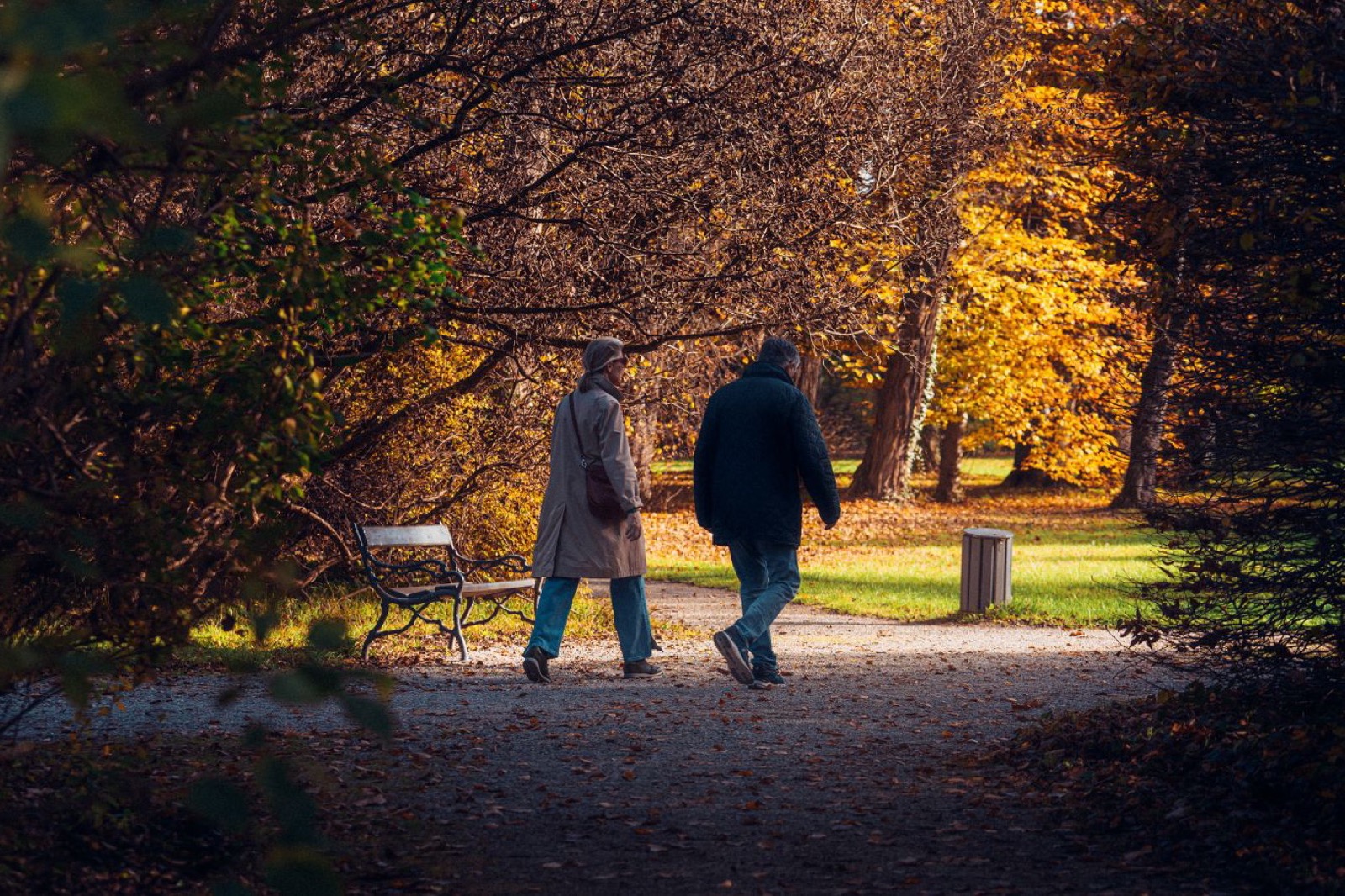 two older adults walking away along a gravel path in a park with autumn trees and a bench