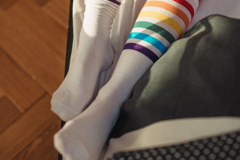 close view of a person sitting on a bed wearing white socks, one with rainbow stripes at the top