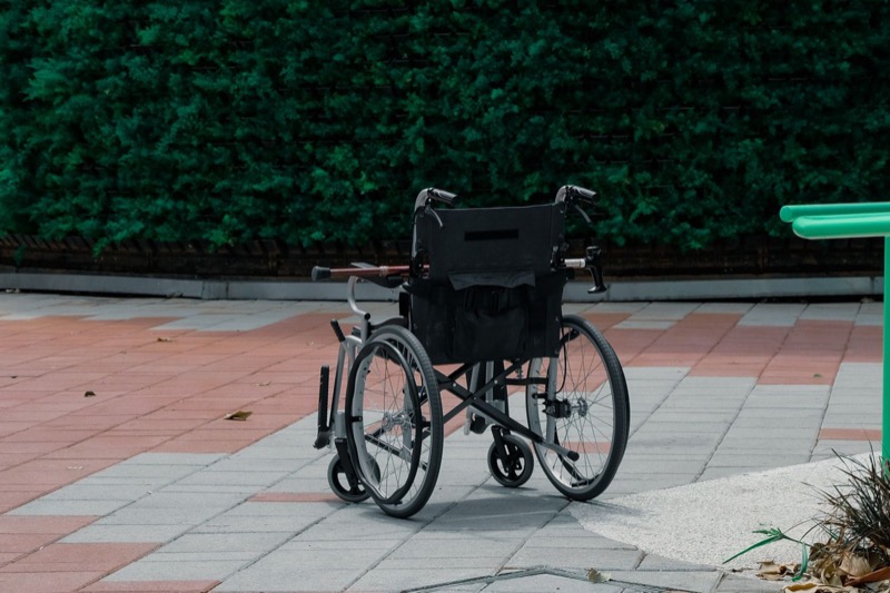 empty black folding wheelchair on a paved brick path in front of a dense green hedge