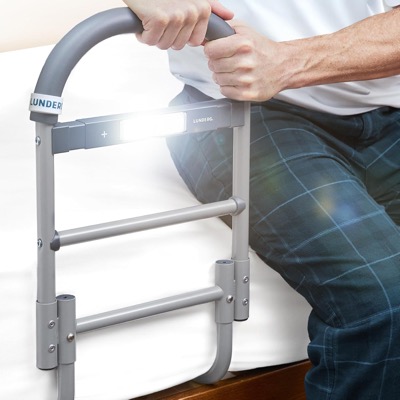 gray steel bed assist rail with a handle and four crossbars attached beside a bed, with a small light